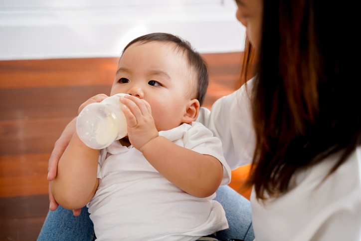 Close-up of Asian mother babysitting and feeding her own son with milk bottle in living room at home. - Motherhood and formula-fed newborn concept